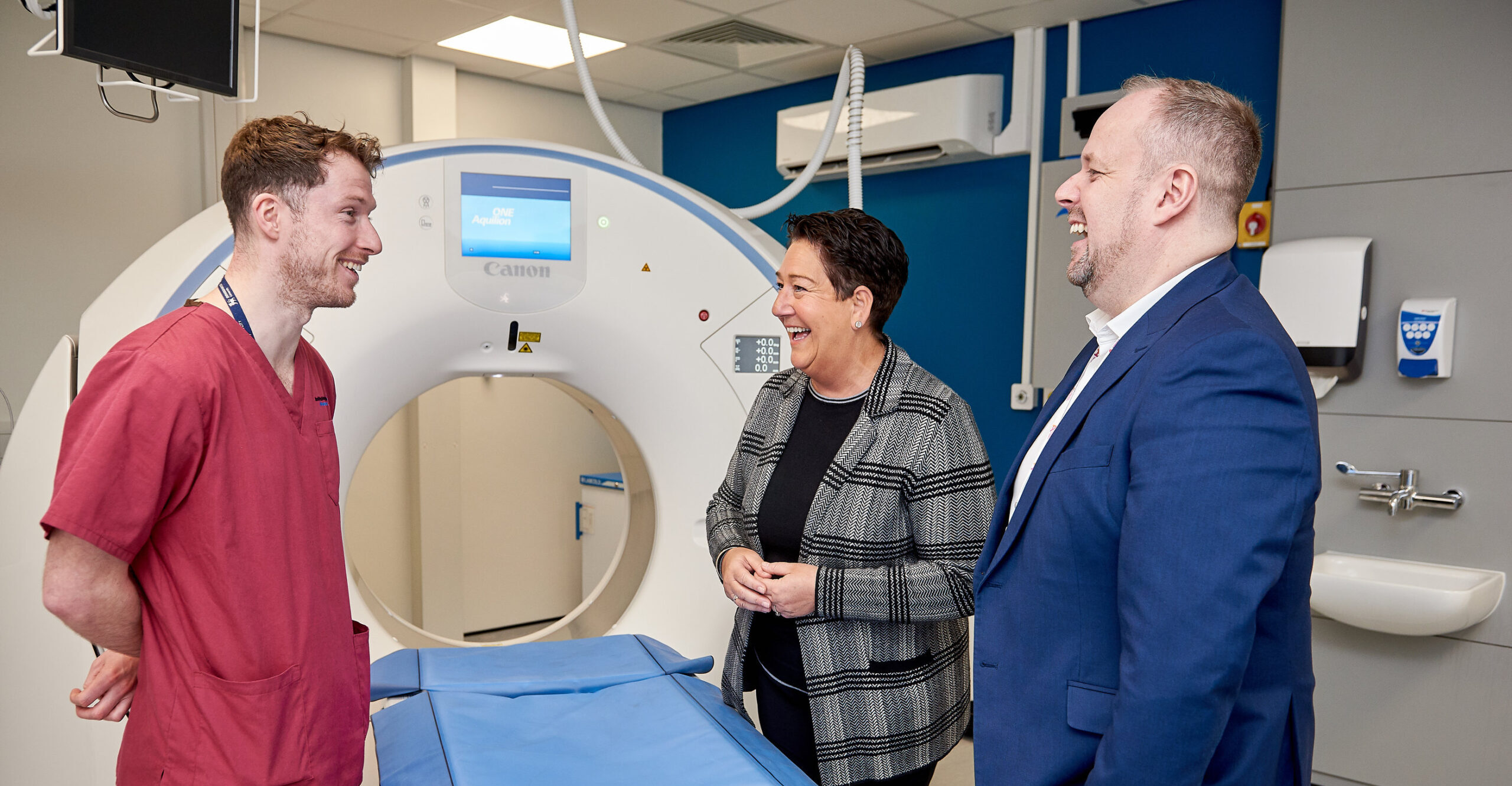 Picture of a CT scanner. Mel Pickup and David Crampsey are standing around the scanner laughing with a radiographer.
