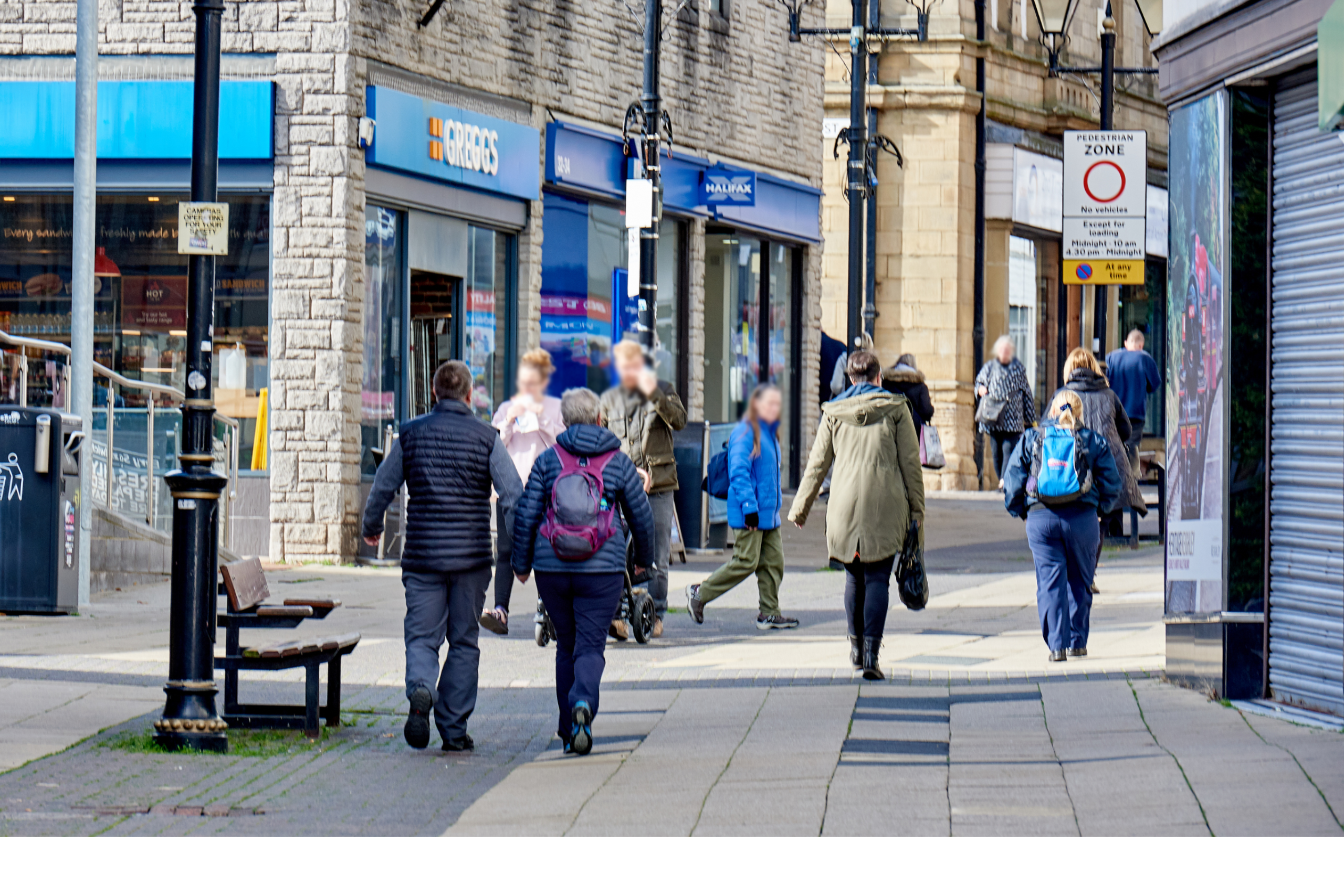 A highstreet with people walking on it. There is a greggs and other shops in the background