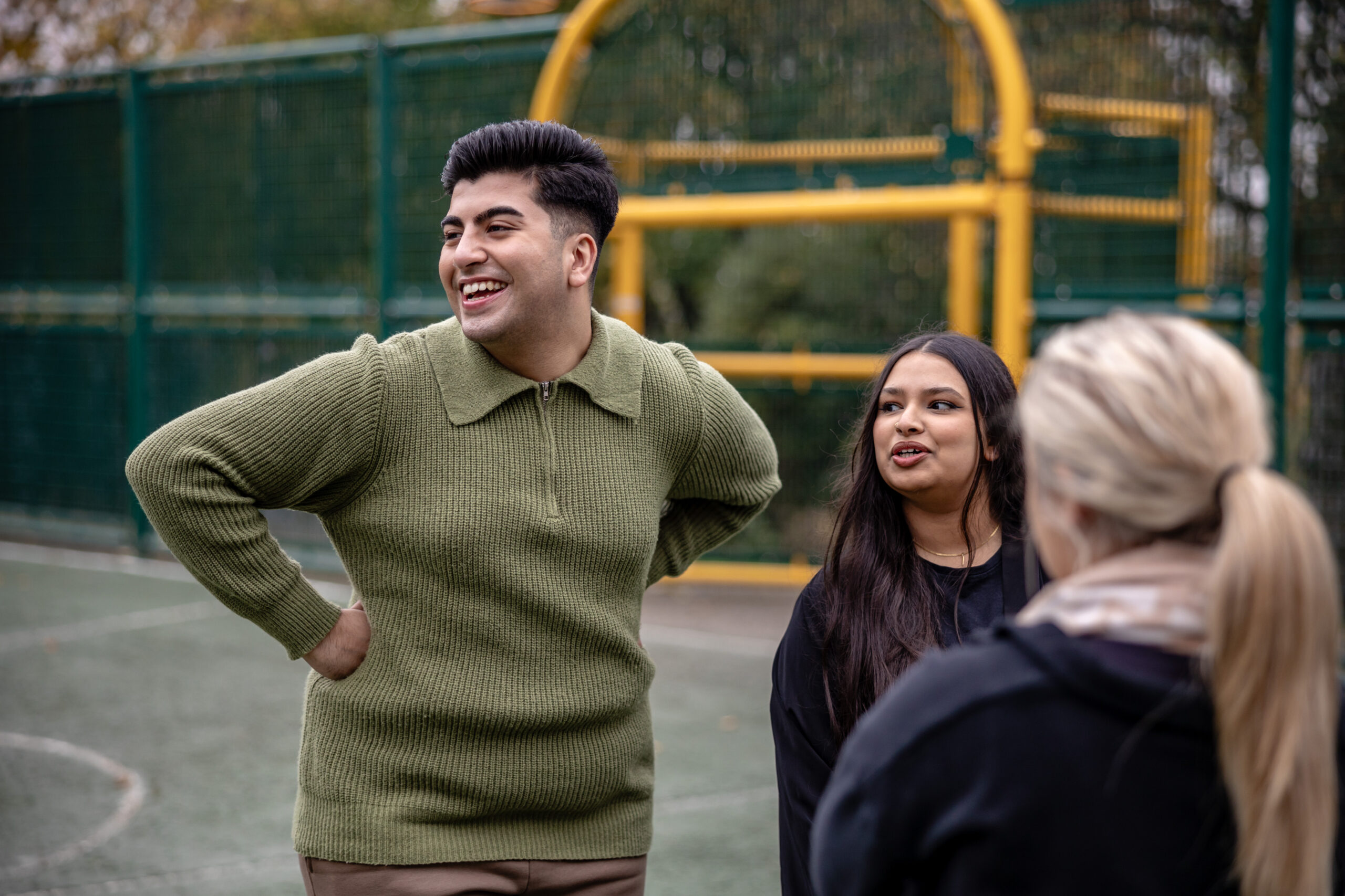 Three young people laughing in a basketball court. Two of them are looking to the left, and the third has their back to the camera.