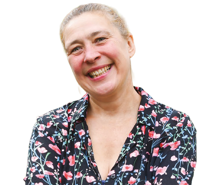 Smiling woman in a floral shirt standing in front of a brick wall with greenery and flowers.