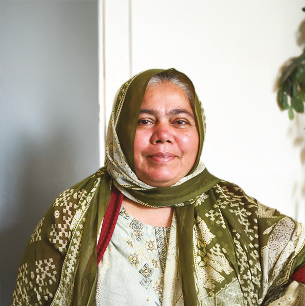 Smiling woman wearing a patterned green and cream traditional outfit with a matching headscarf, seated against a light background.
