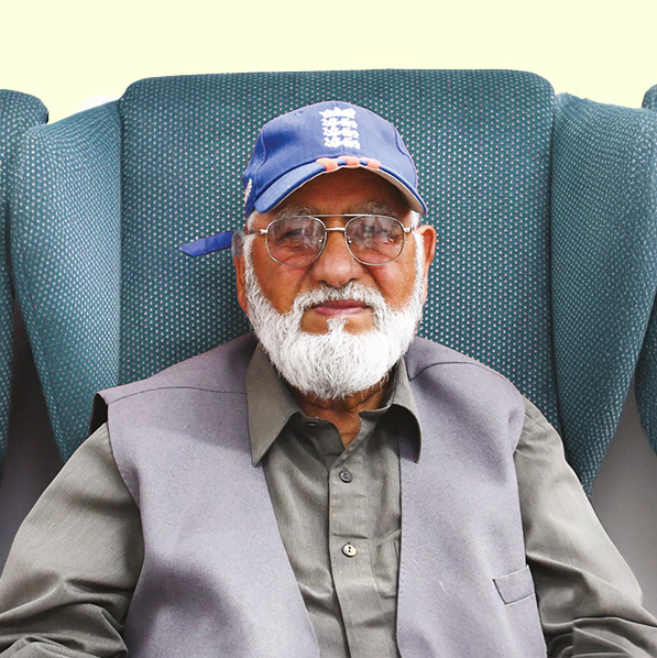 Older man with a white beard wearing glasses, a blue England cricket cap, and a grey waistcoat, smiling while seated against a teal chair.