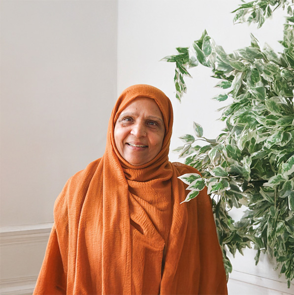 Smiling woman wearing an orange headscarf, standing indoors beside a large green leafy plant.