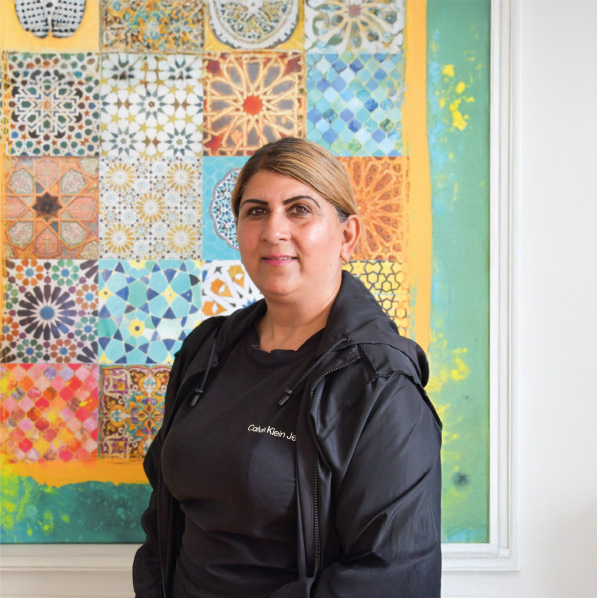 Woman wearing a black jacket and T-shirt, standing indoors in front of a colourful mosaic-patterned artwork.