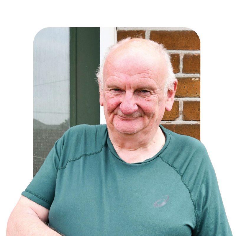 Older man in a green t-shirt smiling outside against a brick wall, sharing his experience with bowel screening.