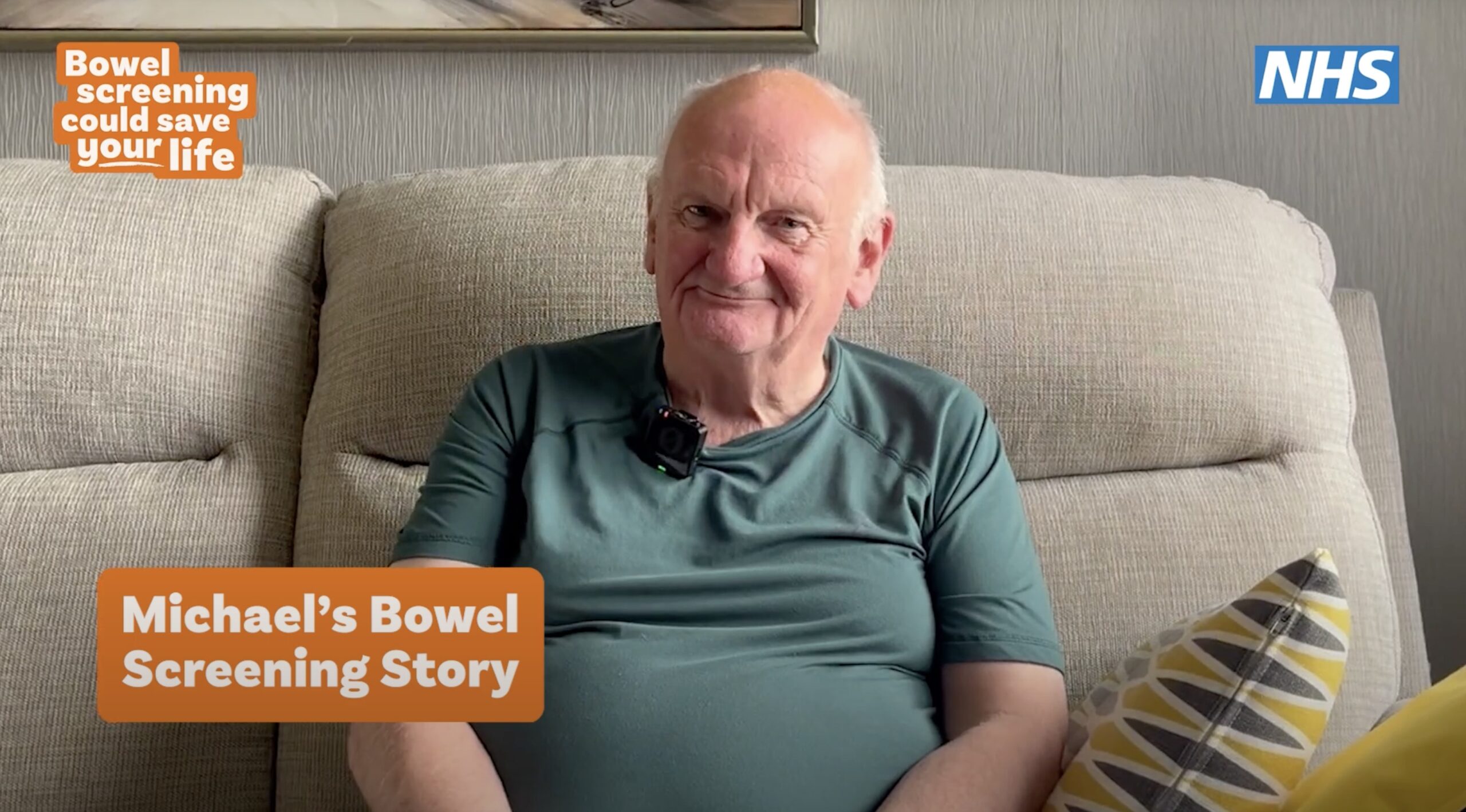 An older man named Michael sits on a beige sofa, smiling at the camera, wearing a green t-shirt with a small microphone clipped on. On-screen text reads: ‘Bowel screening could save your life’ and ‘Michael’s Bowel Screening Story.’ NHS logo in the top right corner.
