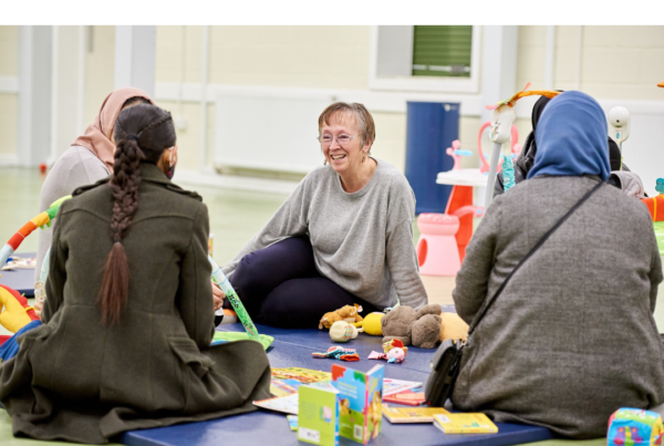 A woman sat on a play mat with toys and other women speaking to her