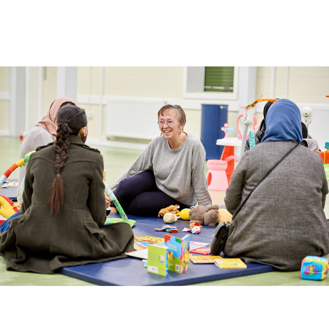 A woman sat on a play mat with toys and other women speaking to her